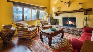 A cozy living room with a fireplace, wooden furniture, and a large window overlooking a scenic view. - Ponderosa Estate | Secluded Luxury (Santa Fe)