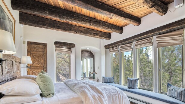 A bedroom with a large bed, wooden beams, a window seat, and a view of trees. - Ponderosa Estate | Secluded Luxury (Santa Fe)