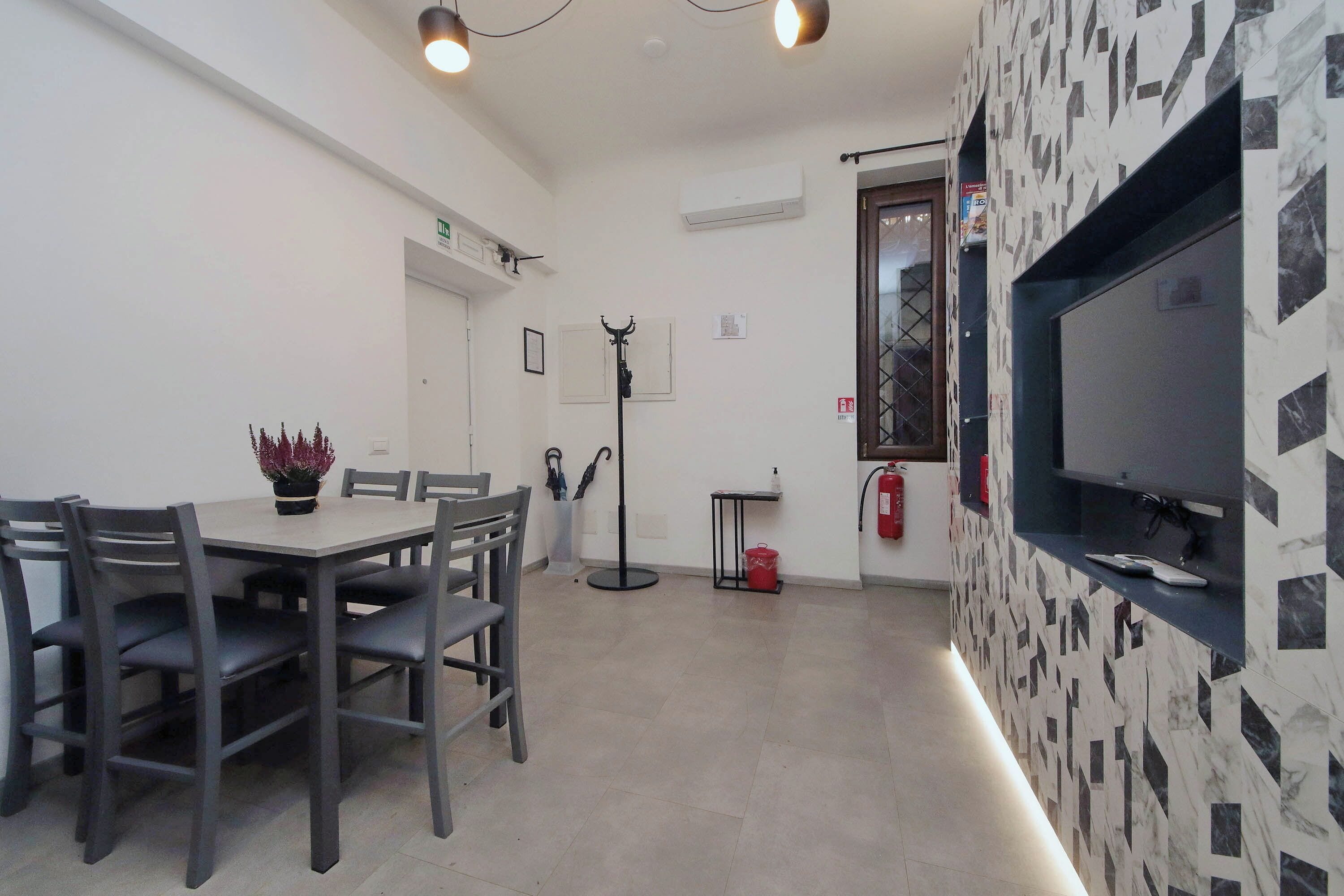 A dining area with a table and chairs, a potted plant, a fire extinguisher, and a television mounted on the wall.