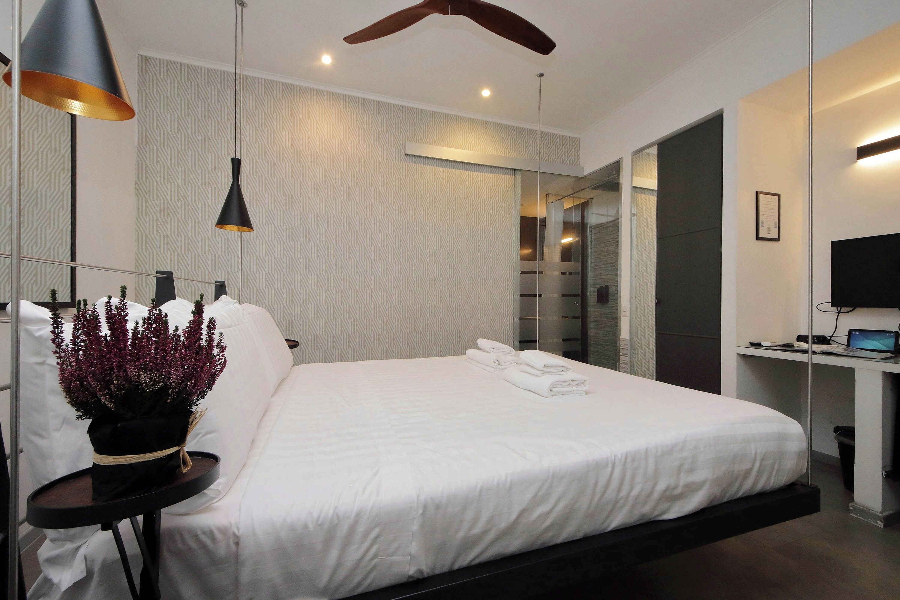 A modern bedroom with a large bed, a small table with a potted plant, a desk with a computer, and a ceiling fan.
