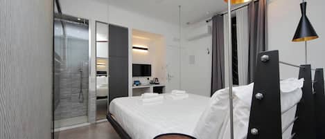 Habitación de hotel moderna con una cama, una mesa redonda, un ventilador de techo y un baño con puerta de vidrio.