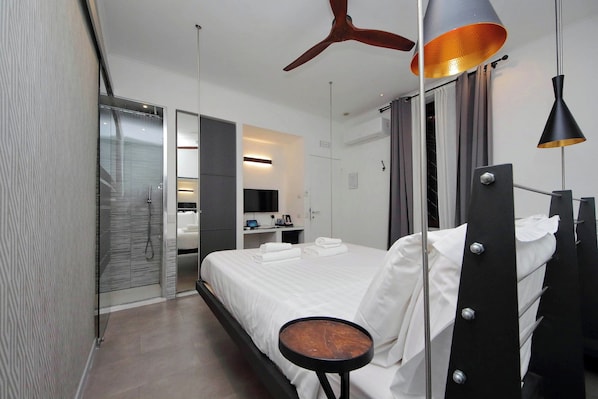 A modern hotel room with a bed, a round table, a ceiling fan, and a bathroom with a glass door.