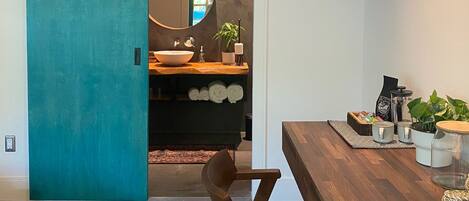 A modern interior with a teal sliding door leading to a bathroom with a large mirror, a sink, and a plant. A wooden dining table with chairs is in the foreground.