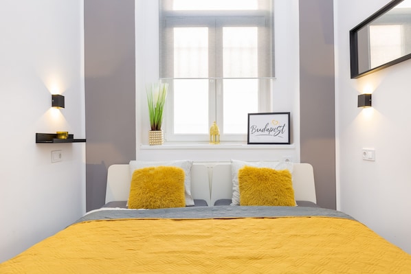 Um quarto moderno com uma cama, dois travesseiros amarelos, uma mesinha de cabeceira com uma planta e um quadro na parede.