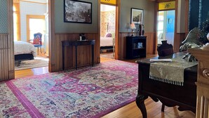 A hotel room with a large bed, a desk, a chair, a small table, a bookshelf, and a large window. - Hotel St. Nicholas (Cripple Creek)