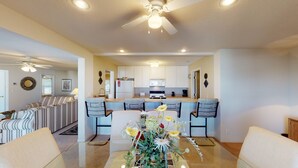 A modern living room with a sofa, a coffee table with flowers, and a dining area with chairs and a kitchen in the background. - Regatta Bay 488 - Walk In 3 Bdrm Condo (Village of Four Seasons)