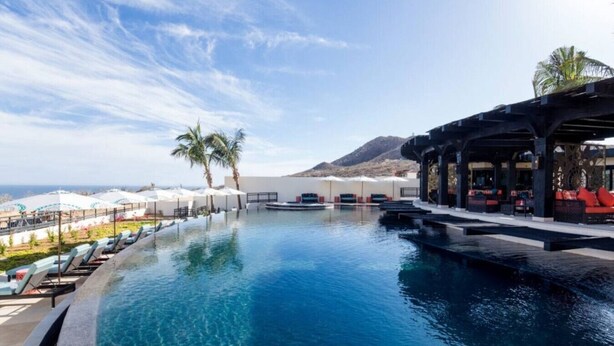 Une piscine avec des chaises longues et des parasols, un espace bar avec des sièges, et une vue sur l’océan et les montagnes.