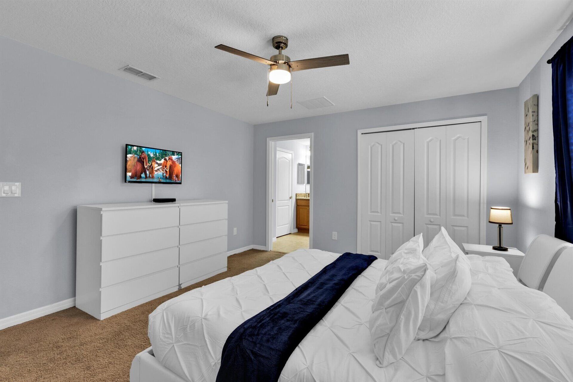 Una camera da letto con un letto, una ventola a soffitto, una TV, un comò e una porta che dà su un'altra stanza.