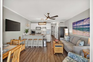 Una cocina moderna con una isla central, taburetes de bar, una mesa de comedor y una sala de estar con un sofá y un cuadro.