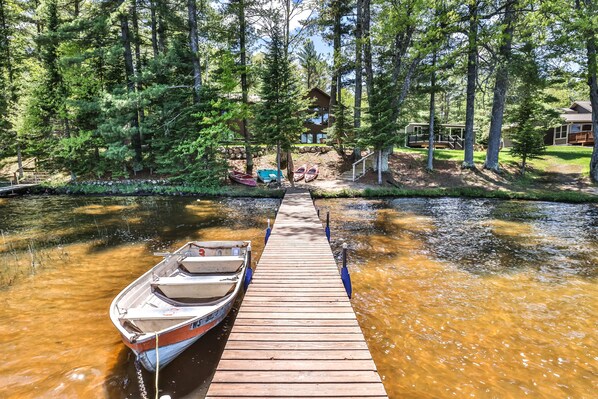 Un muelle de madera que conduce a un pequeño bote, rodeado de árboles y una cabaña al fondo.