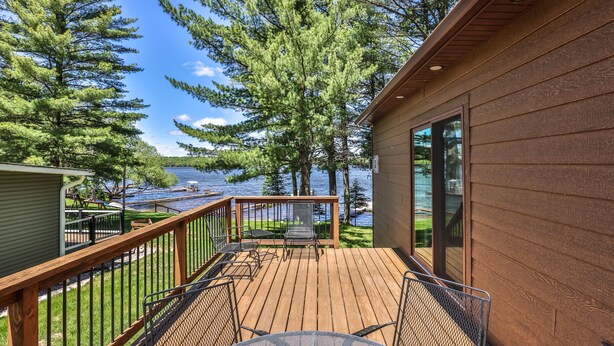 A wooden deck with a table and chairs, overlooking a lake with boats and surrounded by trees.