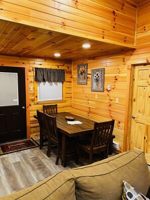 A cozy cabin interior with a wooden dining table, four chairs, a sofa, a window with curtains, and two wall-mounted pictures. - RIVERSIDE RENOVATED 1950's schoolhouse, HOT TUB, Pellet Stove, Pet Friendly (Elkins)
