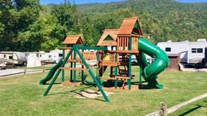 A playground with a slide and climbing structures, surrounded by grass and trees, with RVs in the background. - Ugly Cabin, Stunning Escape – Hot Tub! Fire Pit! Fast WIFI! Sunroom! Hiking! (Elkins)