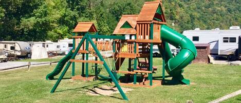 Un parque infantil con tobogán y estructuras para escalar, rodeado de césped y árboles, con vehículos recreativos al fondo.