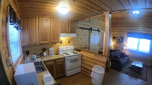 A wooden cabin interior with a kitchen featuring a stove, sink, and a coffee maker. There is a couch and a small table in the living area. - Ugly Cabin, Stunning Escape – Hot Tub! Fire Pit! Fast WIFI! Sunroom! Hiking! (Elkins)
