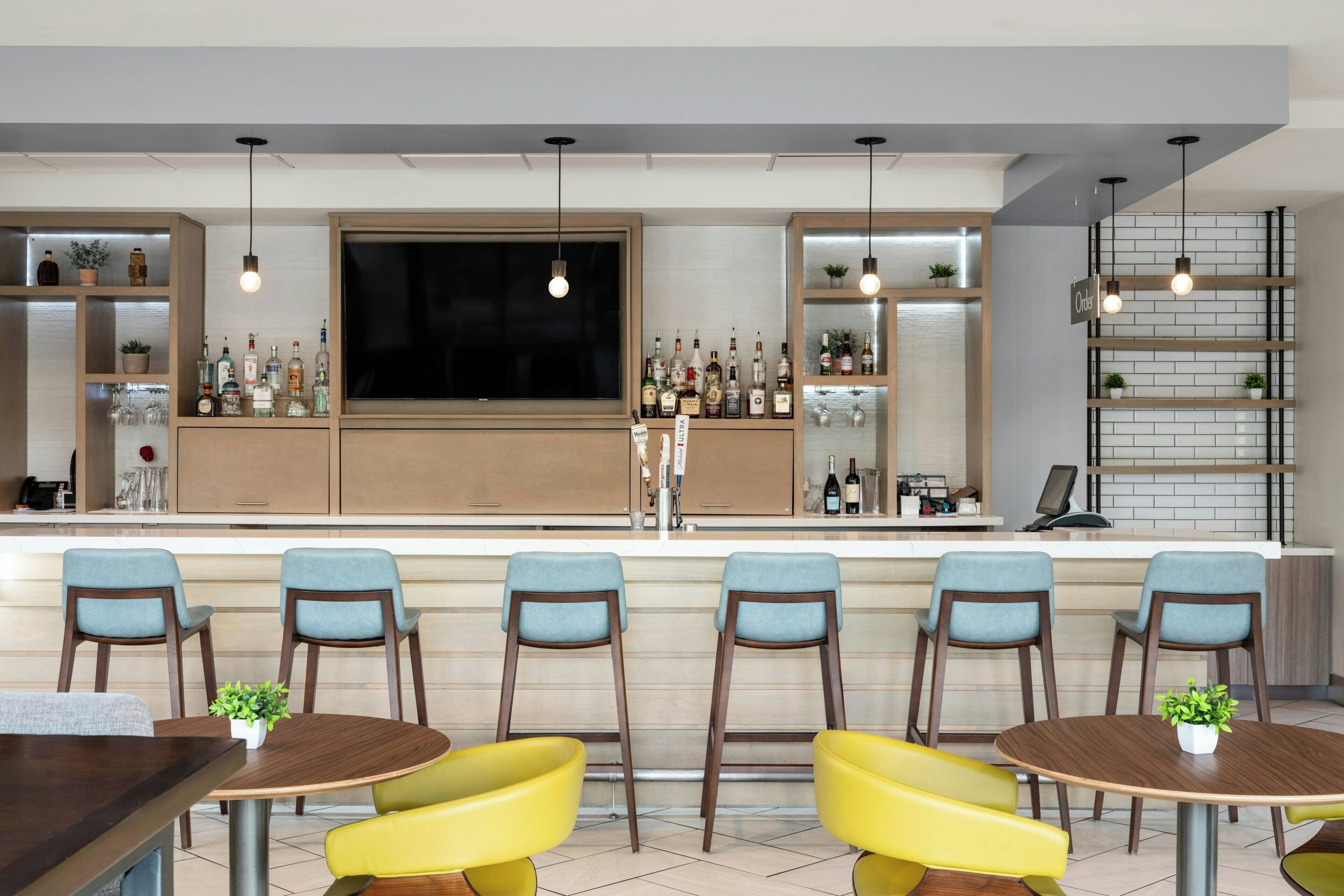 A bar area with multiple seating options, including bar stools and round tables, with a variety of bottles and glasses on shelves.