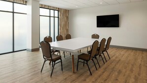 Une salle de conférence avec une table blanche, six chaises, une grande fenêtre avec des rideaux et un téléviseur à écran plat fixé au mur.