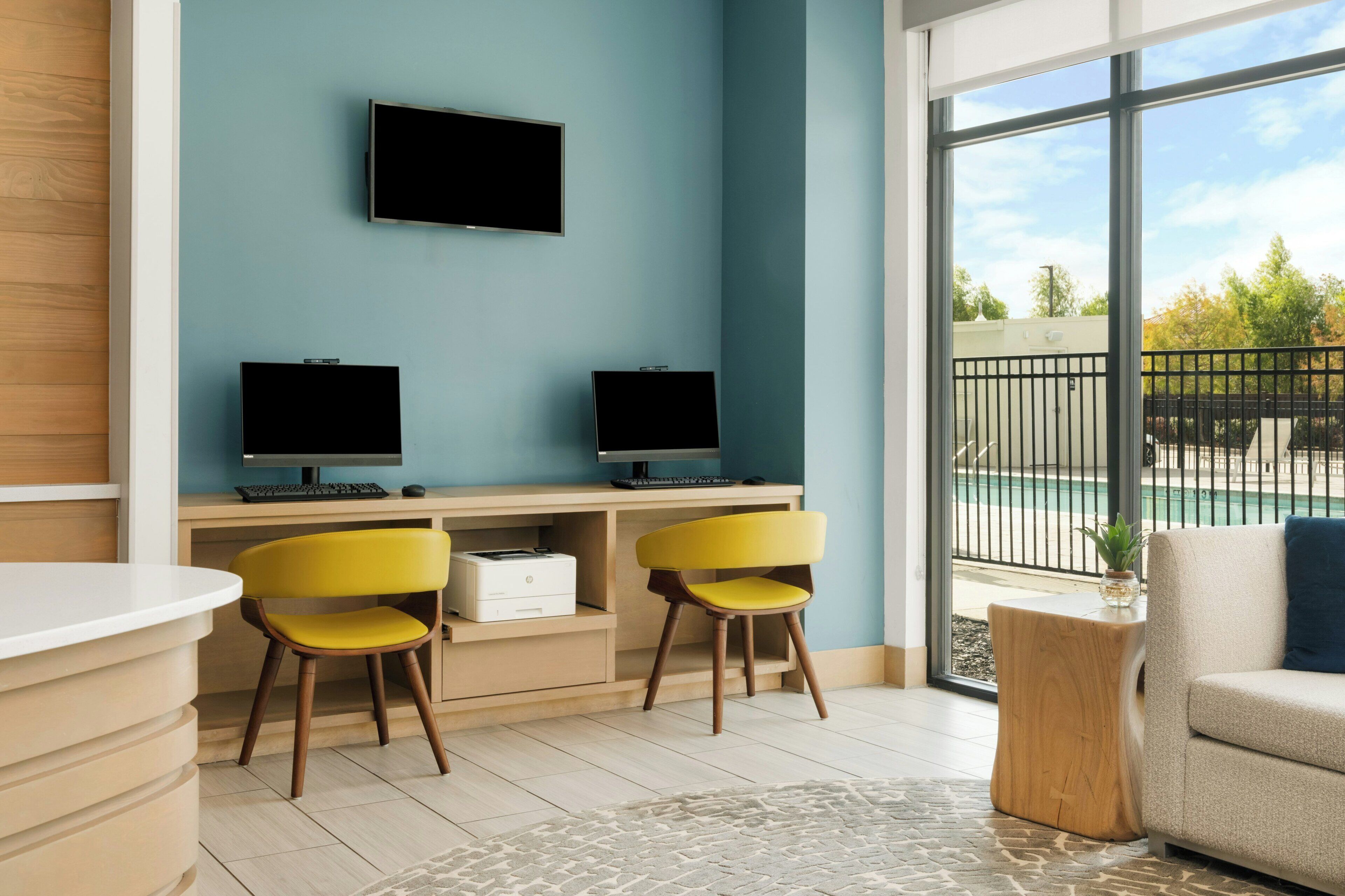 Un espacio de oficina moderno con dos escritorios, dos sillas, un monitor de pantalla plana, una computadora de escritorio, una impresora y vistas a una piscina a través de amplias ventanas.