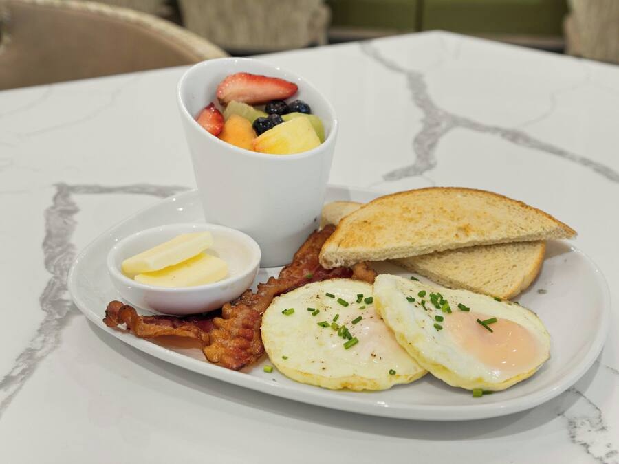 A breakfast plate with eggs, bacon, toast, butter, and a fruit cup.