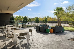 Patio esterno con tavoli e sedie, piscina e area recintata con alberi e cielo sereno.