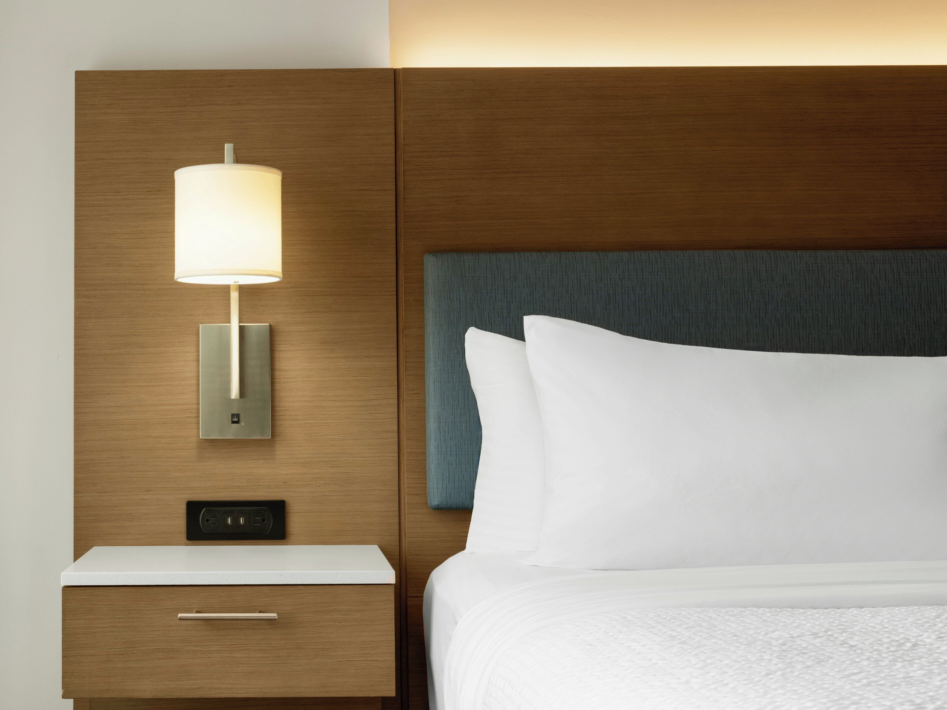 A hotel room with a wooden headboard, a bedside lamp, a nightstand with a drawer, and a bed with white bedding.