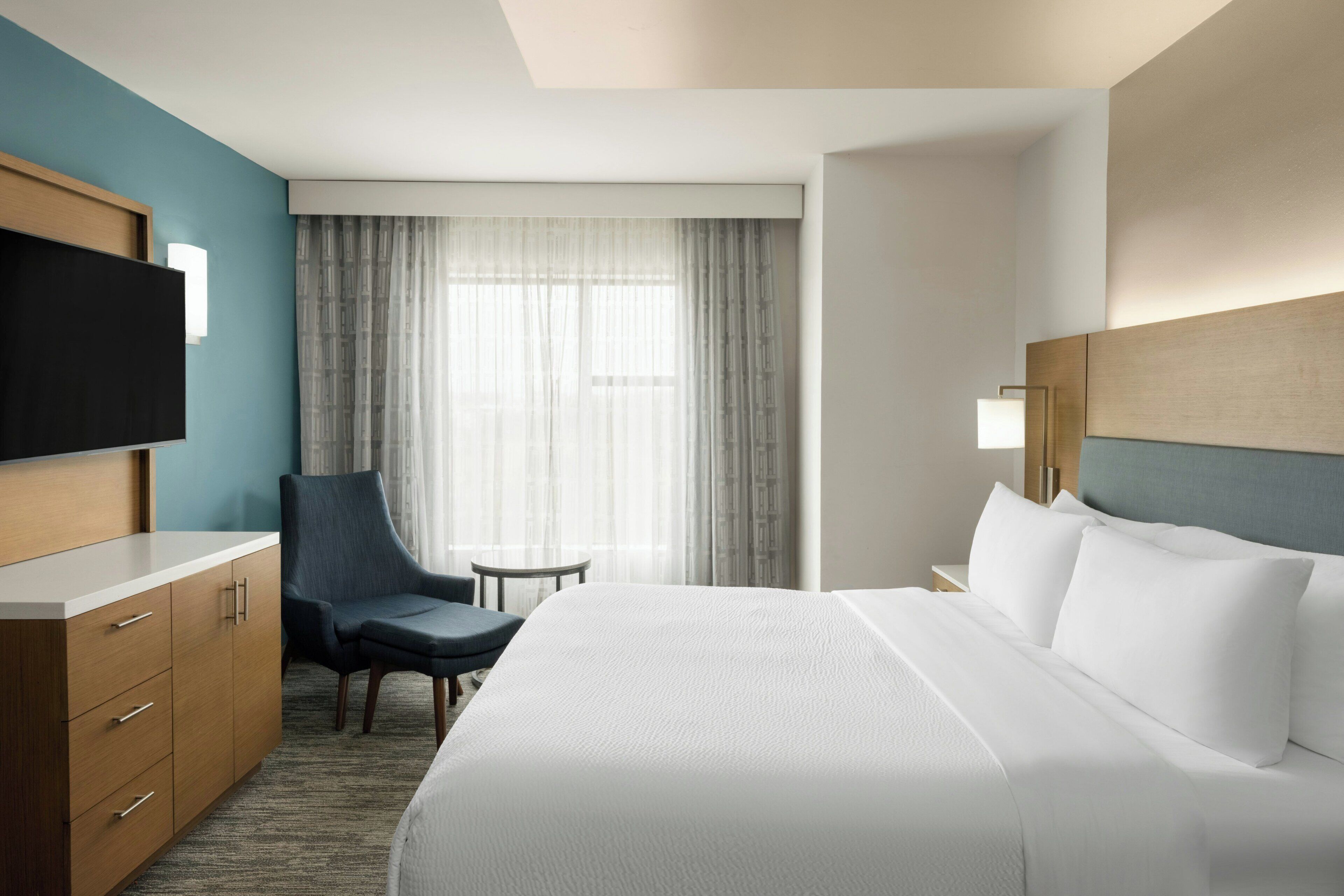 A hotel room with a bed, a desk, a chair, a TV, and a window with curtains.
