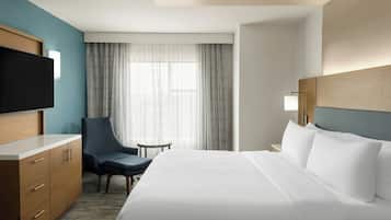 Quarto de hotel com uma cama, escrivaninha, cadeira, televisão e janela com cortinas.