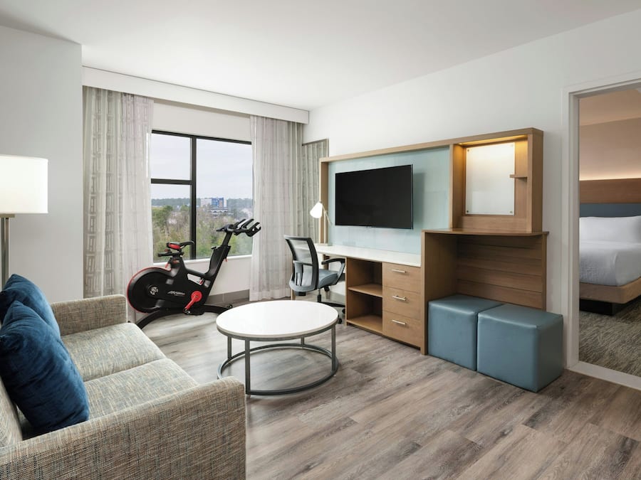 A hotel room with a bed, a desk, a chair, a TV, a treadmill, and a view of the city.