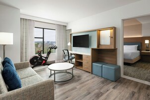 Habitación de hotel con cama, escritorio, silla, televisor, cinta de correr y vista de la ciudad.