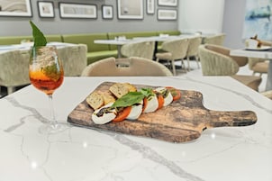 Una mesa de comedor con una tabla de madera que sostiene un aperitivo de tomate y mozzarella, acompañado de un vaso de bebida roja.