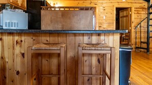 Ein Holzhütten-Interieur mit Küchenbereich, zwei Barhocker und einer Treppe, die zu einer oberen Ebene führt.