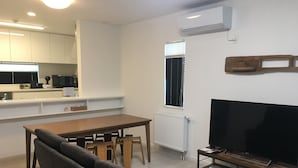 Un salon moderne avec une table à manger en bois, un canapé, un meuble pour la télévision et un coin cuisine doté de meubles blancs et d’un micro-ondes encastré.