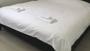 Un lit bien fait, avec du linge de lit et des serviettes blanches, une tête de lit sombre, une petite table de chevet avec une lampe et un climatiseur fixé au mur.