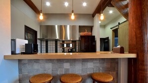 A modern kitchen with a wooden bar counter, three stools, stainless steel appliances, and hanging light fixtures. - Chise Fukuro (Furano)