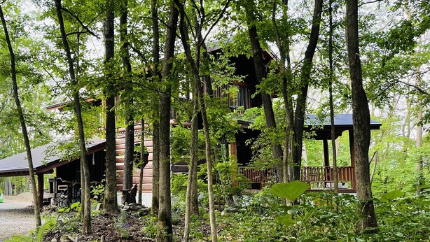 A rustic cabin surrounded by tall trees, with a wooden deck and a covered porch. - Chise Fukuro (Furano)