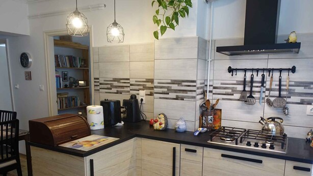 A modern kitchen with a central island, wooden cabinets, a gas stove, hanging kitchen utensils, and a bookshelf in the background. - Holiday home with garden and terrace near the center (Siófok)