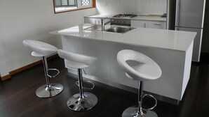 A modern kitchen with white cabinetry, a stainless steel sink, a gas stove, and a refrigerator. There are three white bar stools with chrome bases. - Furano Lodge 10 (Furano)