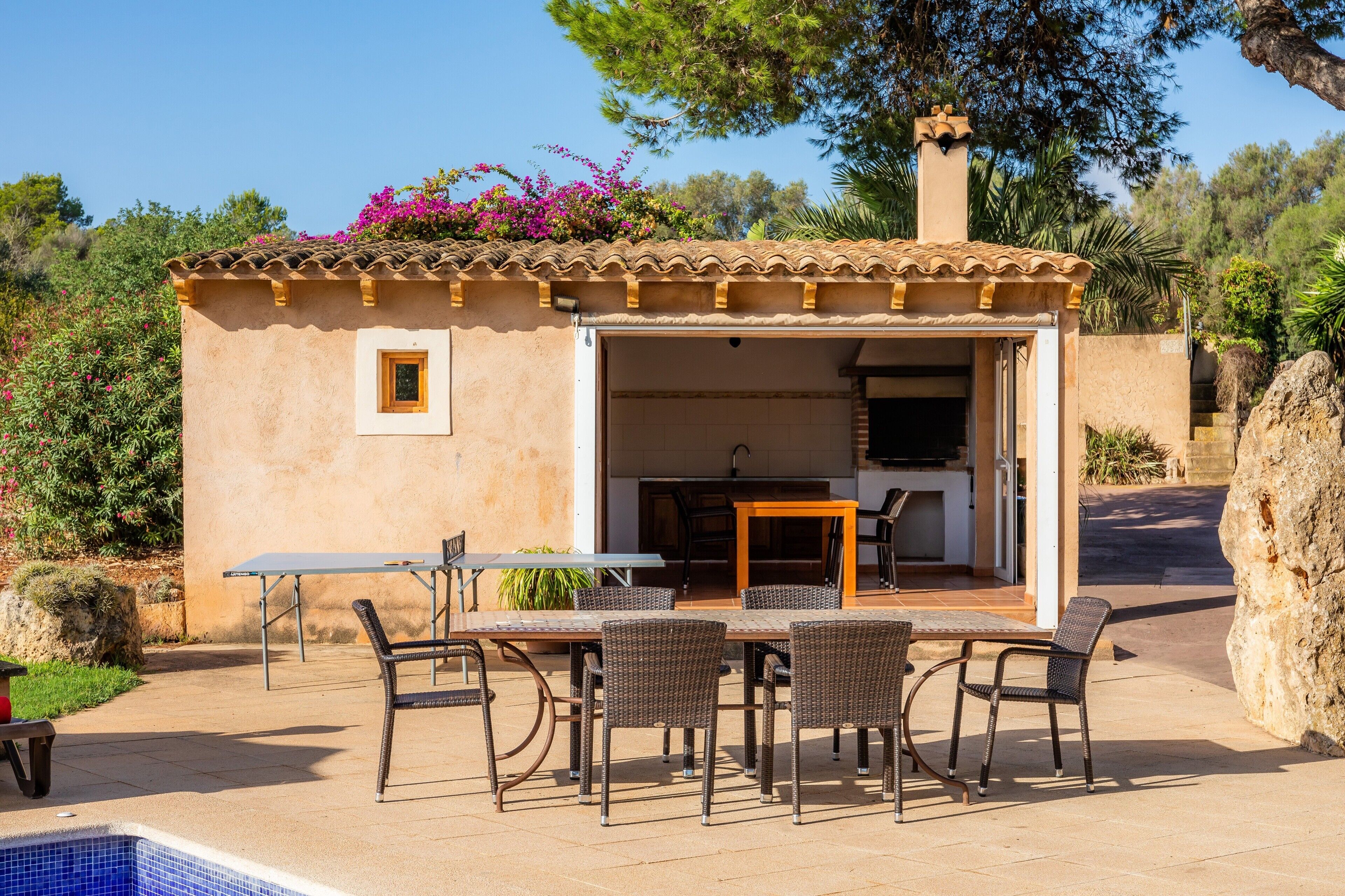 Un pequeño espacio exterior con cocina, mesa, sillas, piscina y una pared de piedra.