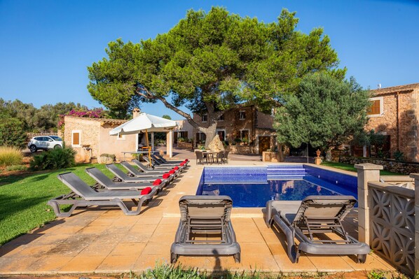 A pool area with lounge chairs, a stone building, and a large tree.