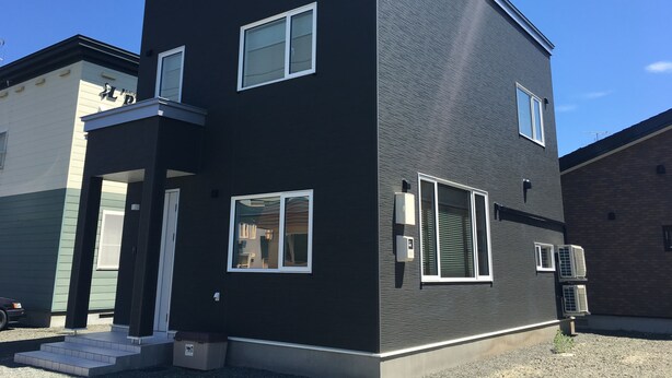 A modern two-story building with a dark exterior, white-framed windows, and a visible air conditioning unit. - Villa Paolo (Furano)
