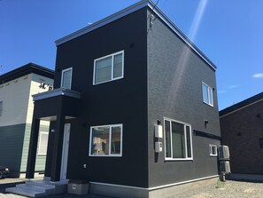 Un edificio moderno de dos pisos con fachada oscura, ventanas con marcos blancos y una unidad de aire acondicionado a la vista.