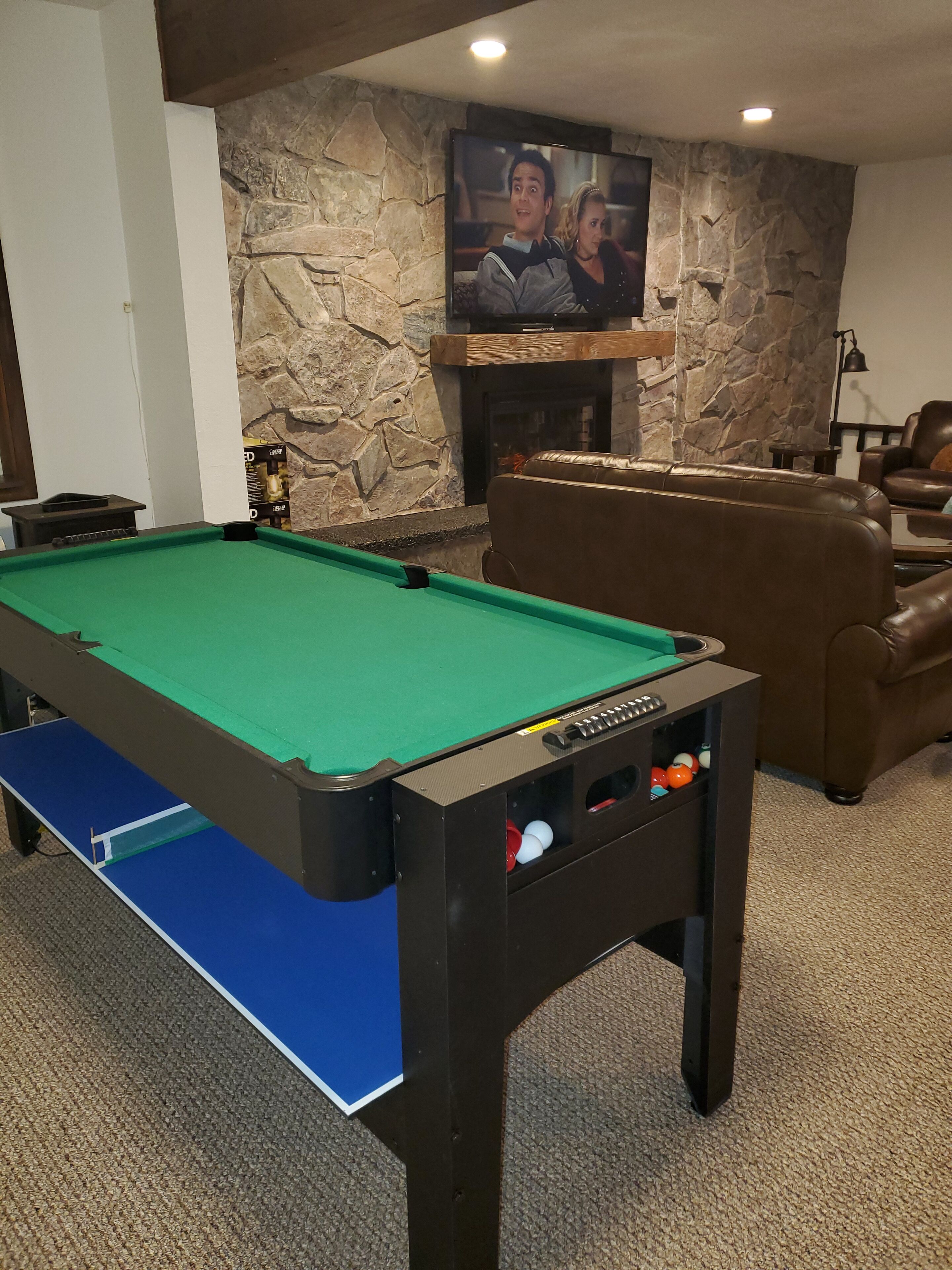 A pool table with a blue felt table and a green felt playing surface, a stone wall with a fireplace, and leather sofas in a room with carpet flooring.