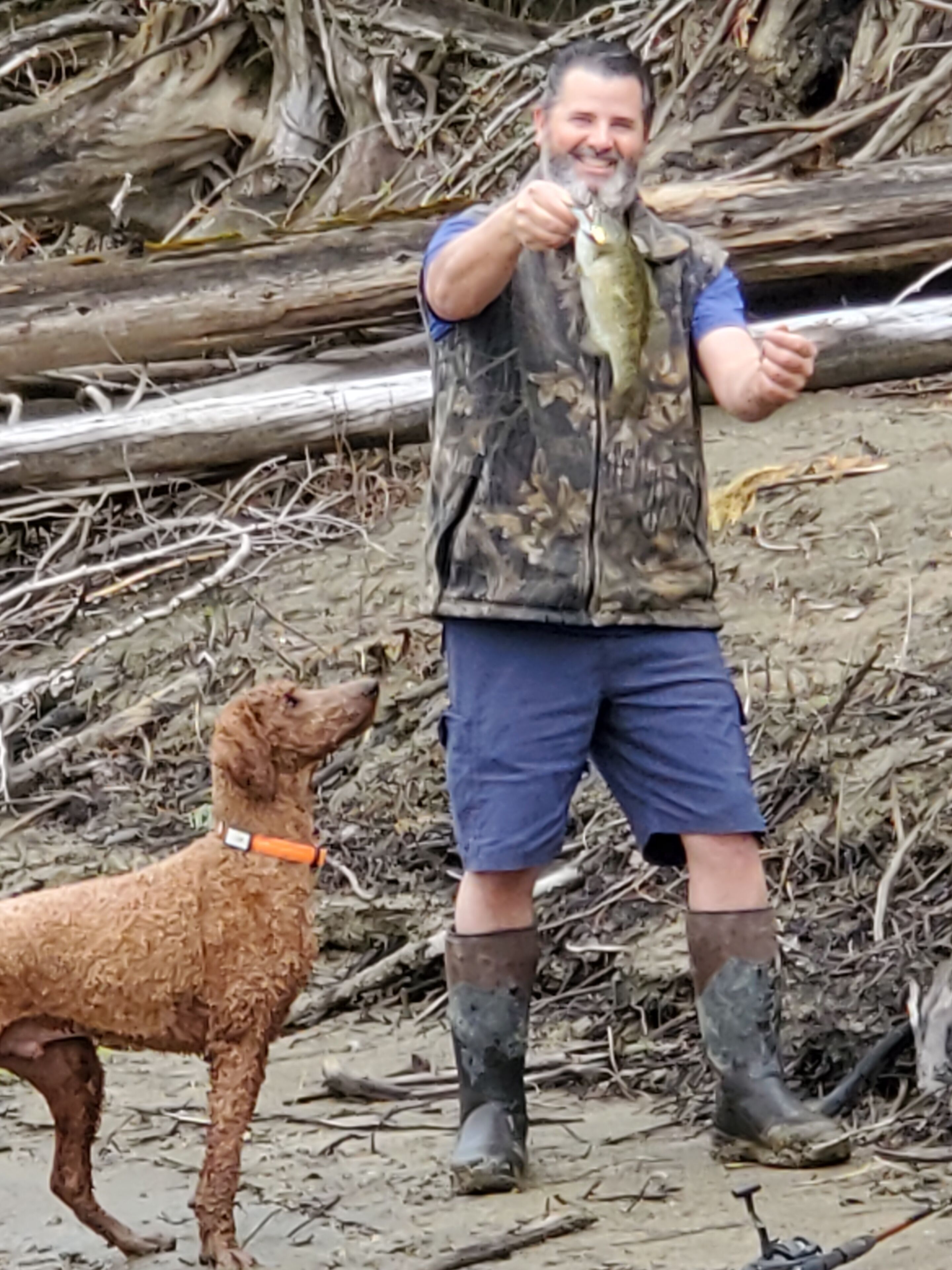 A person holding a fish, standing on a beach with a dog nearby.