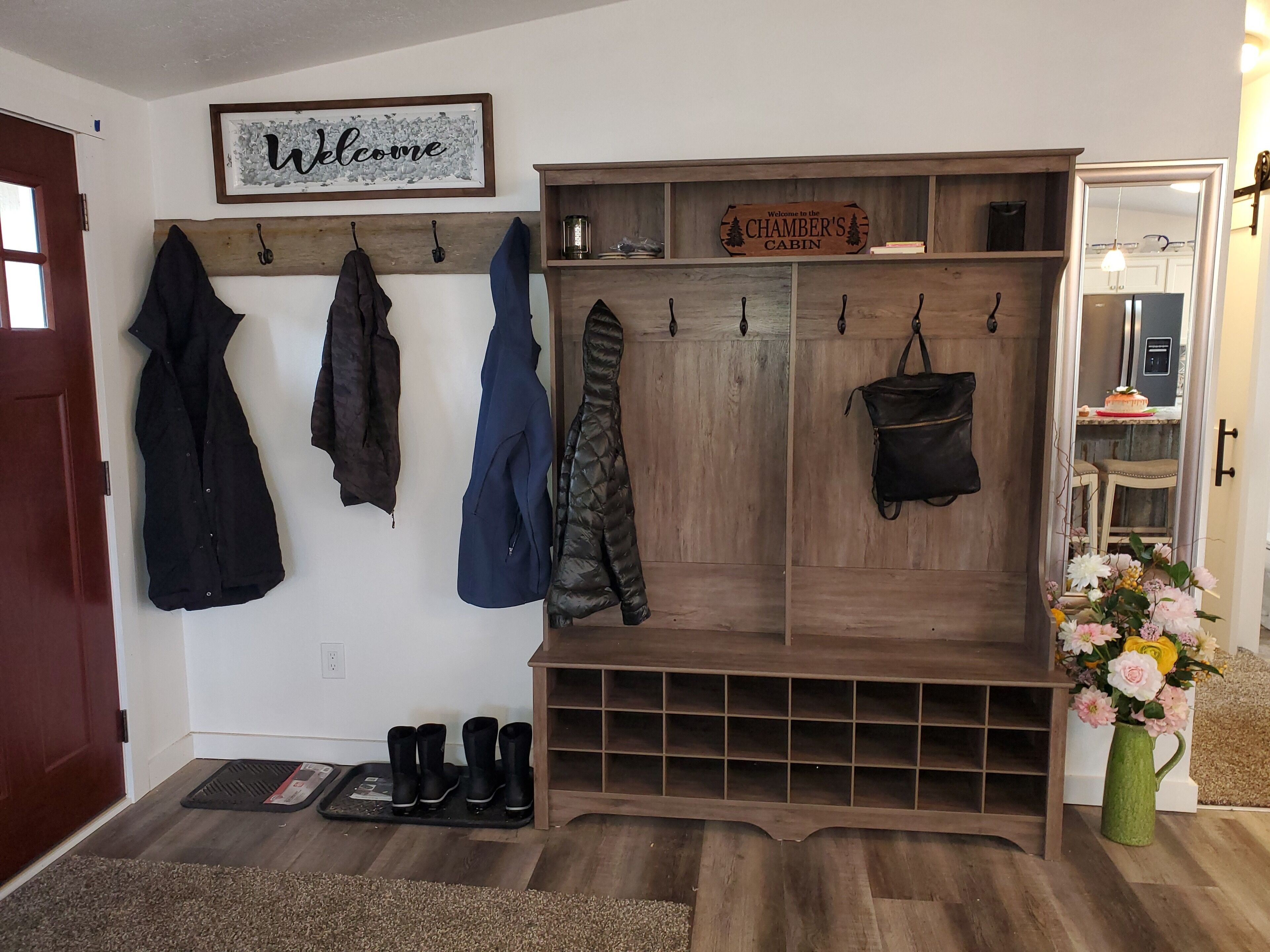 Un perchero de madera con abrigos y un letrero de bienvenida, un mueble de madera con un cartel que dice "Welcome to the CHAMBER'S CABIN", y un jarrón verde con flores.