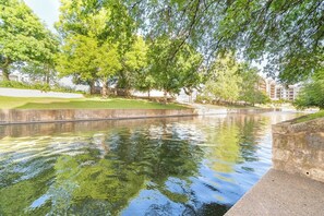 A calm water feature with trees and a concrete edge, surrounded by a grassy area and buildings in the background. - San Antonio | Friends & Family River Walk Trip | Pool + Downtown | 4BR Sleeps 10 (San Antonio)