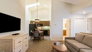 Ett modernt hotellrum med en platt-TV, en byrå i trä, en matplats med bord och stolar, en soffa och ett kök i bakgrunden.