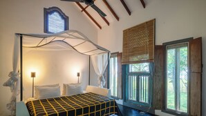 A bedroom with a bed under a canopy, a bedside table, a bench, a standing lamp, and a window with a view of greenery.
