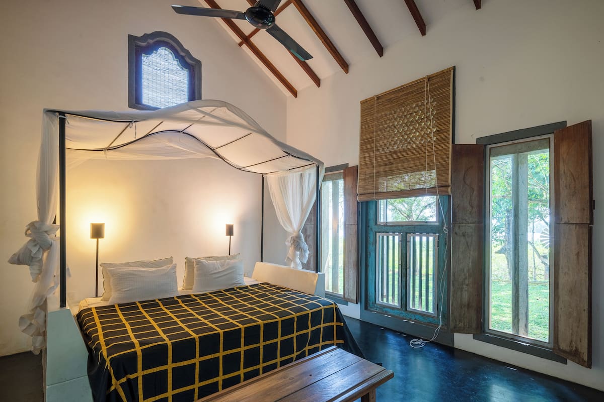 Een slaapkamer met een bed onder een hemelbed, een nachtkastje, een bankje, een staande lamp en een raam met uitzicht op groen.
