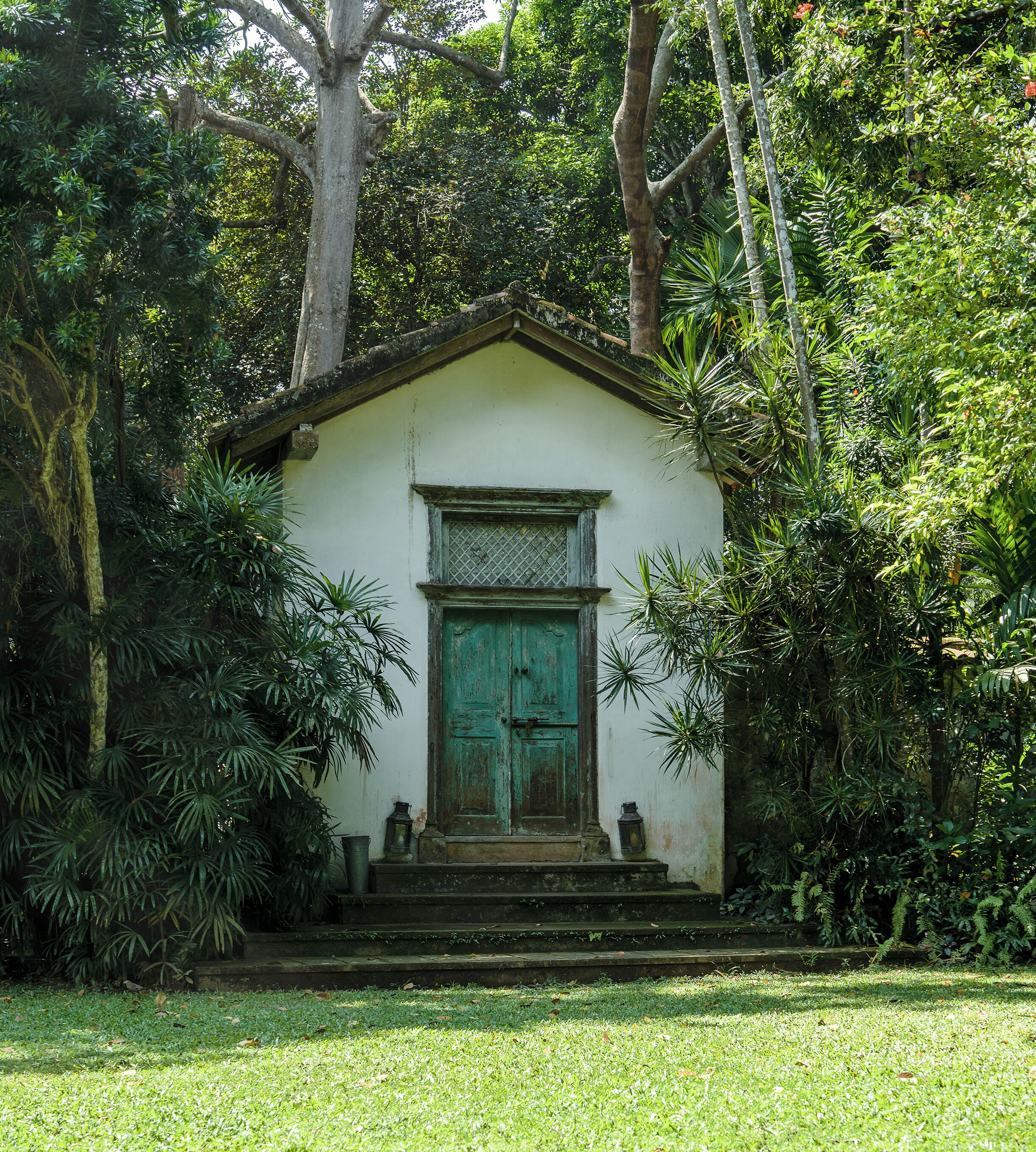 Um pequeno edifício branco com porta verde, rodeado por vegetação exuberante.