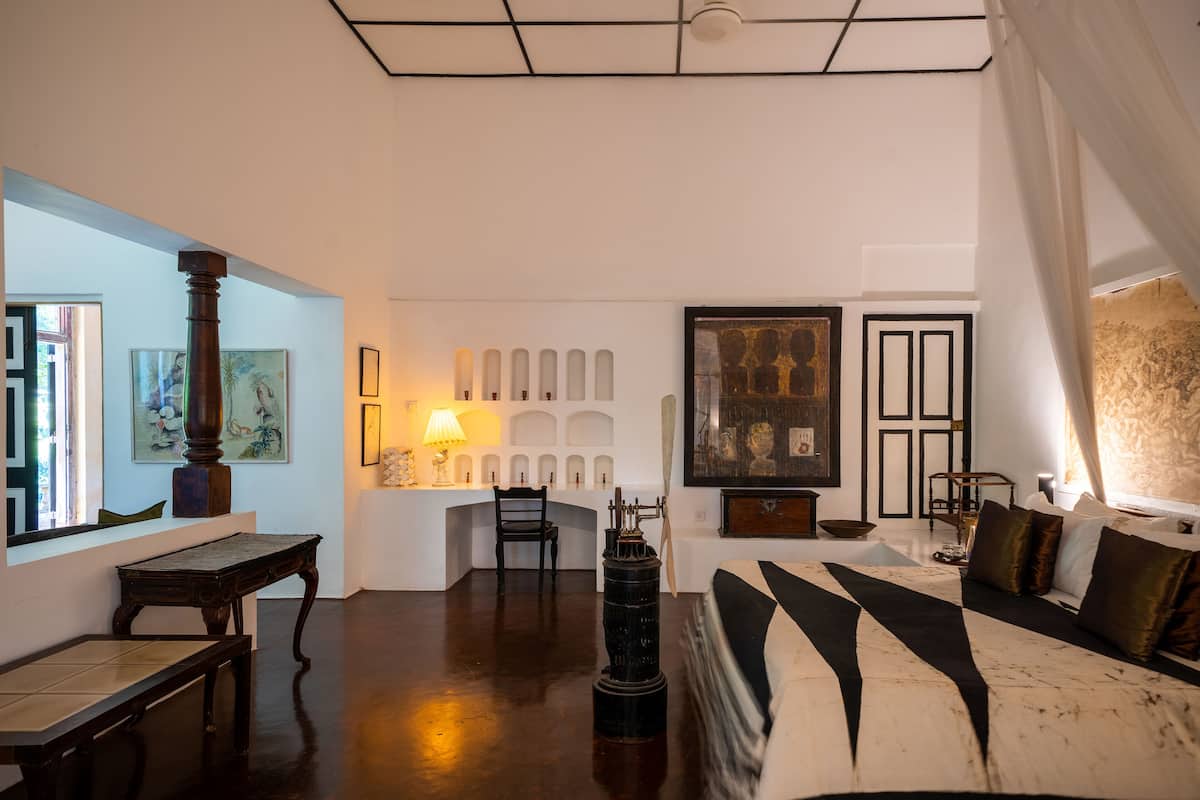 Ruime kamer met een groot bed, een houten tafel, een stoel en een bureau. Er staat een lamp op het bureau en een schilderij hangt aan de muur.